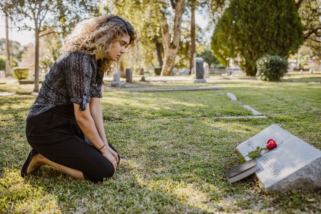 Woman kneeling beside a gravestone with a red rose, in a peaceful cemetery setting