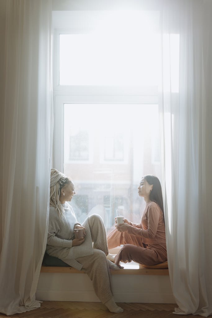 Two women sitting on a window seat, talking and holding mugs, in soft natural light — representing the personal inspiration behind a digital memorial service in Edinburgh.