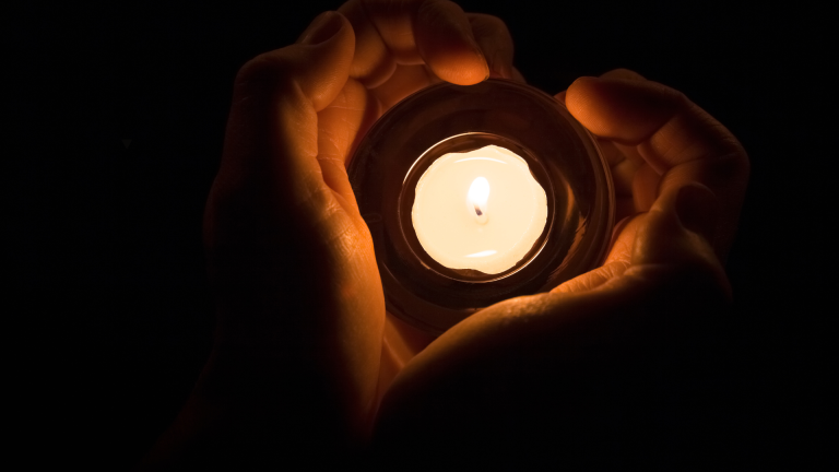 Hands gently holding a lit candle in the dark, symbolising remembrance and digital memorials