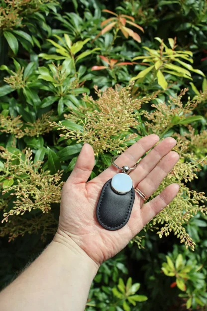 Unmarked leather NFC keychain for a digital memorial, held in hand with green plants in the background to show scale.