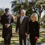 wo men dressed formally at a cemetery during a memorial service, standing among gravestones with trees in the background.
