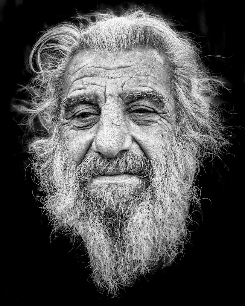 Black and white portrait of a senior man with a prominent beard and expressive features against a dark background.