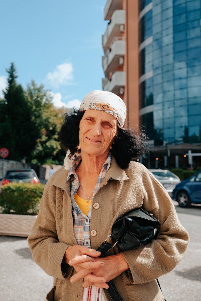 Elderly woman smiling outside in urban setting with headscarf.