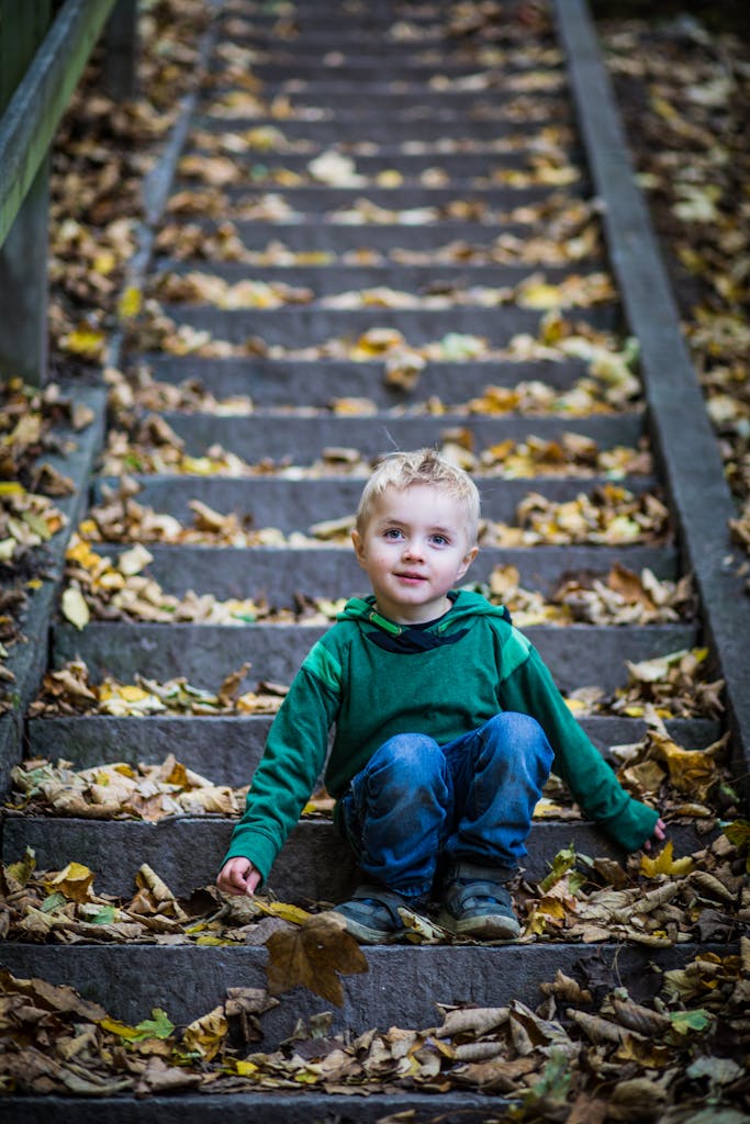 Young boy sitting on outdoor steps covered in autumn leaves, smiling at the camera.